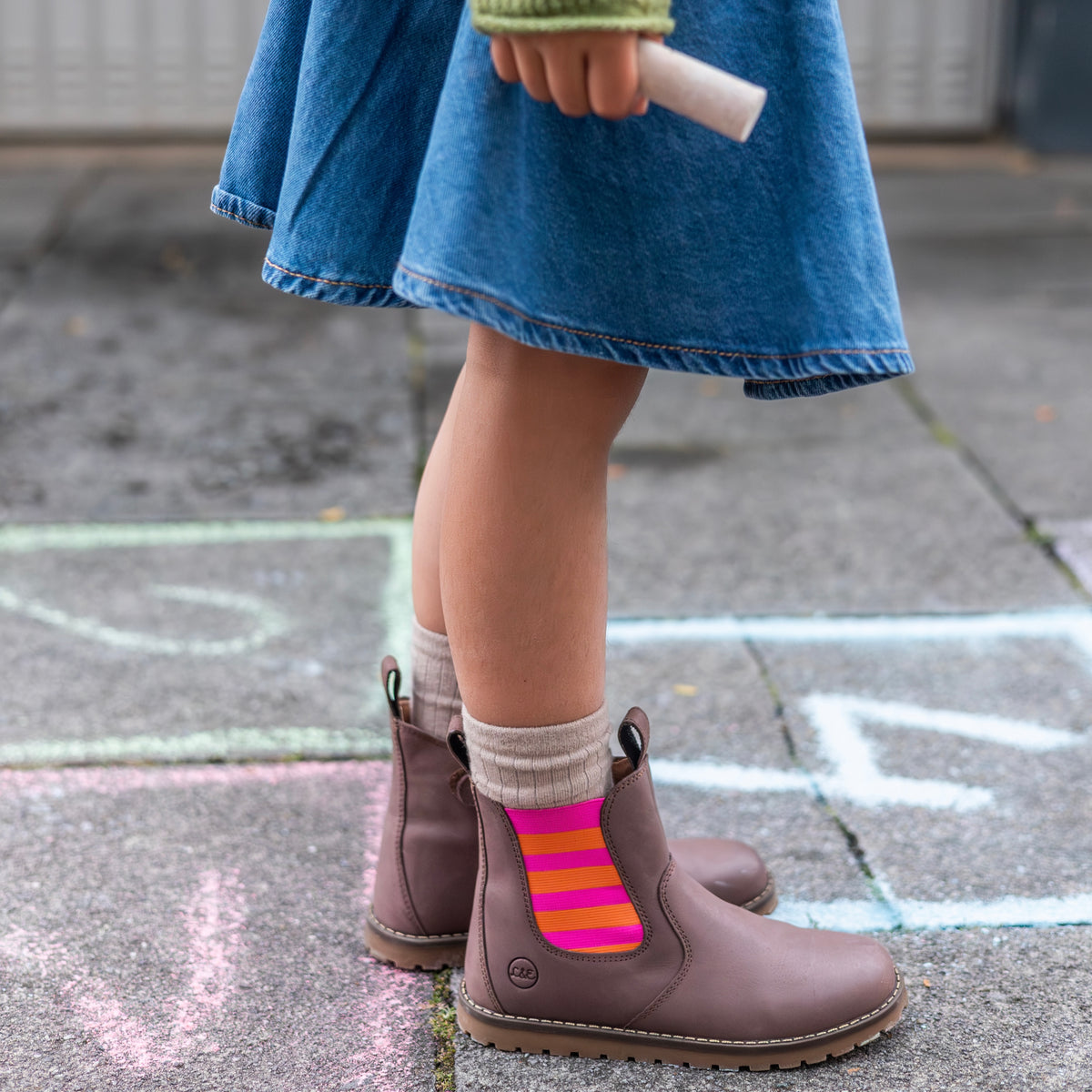 Child wearing the pink and orange Chelsea boots on a sidewalk chalked with hopscotch squares