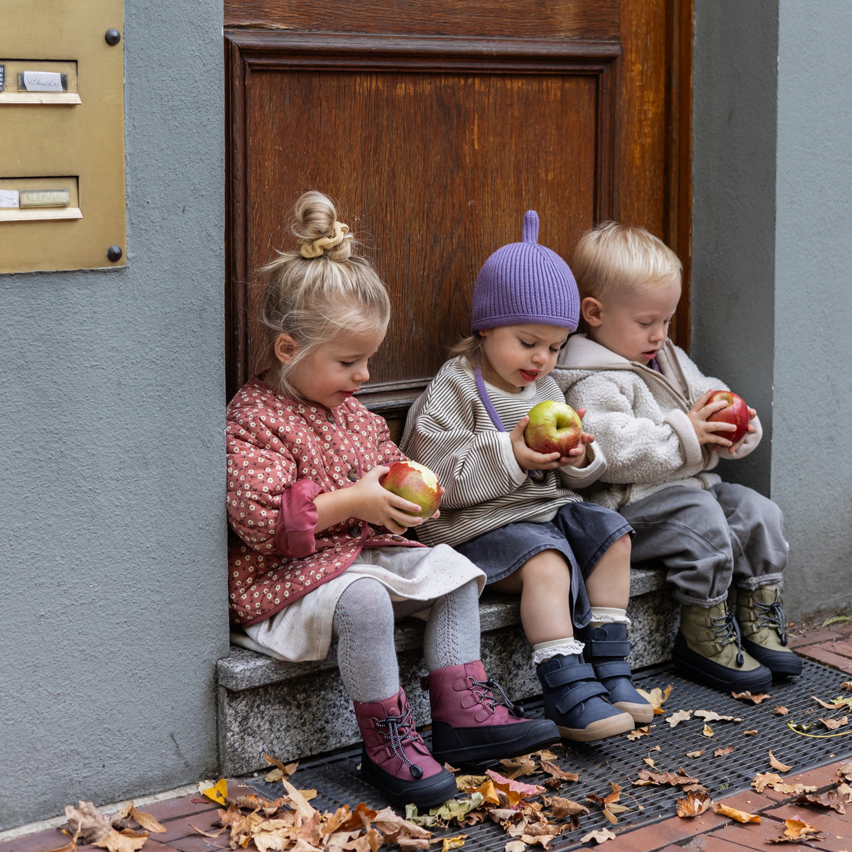 Three children on a doorstep wearing the First Step Finn in berry, navy and khaki